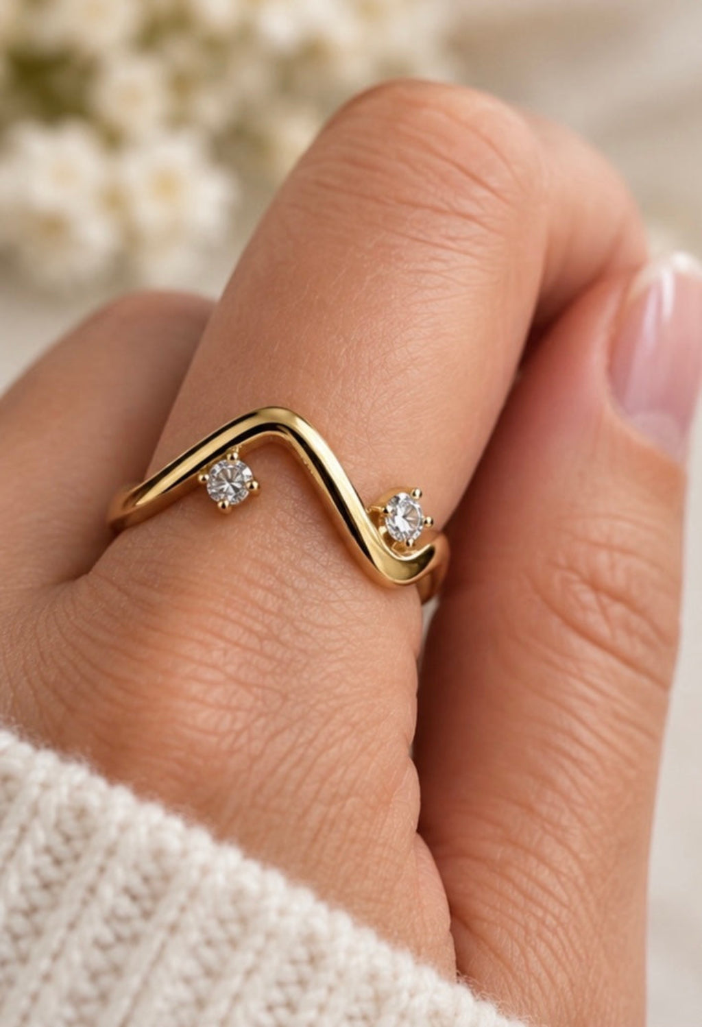 A close-up of a hand wearing a gold wavy ring with two small clear gemstones, set against a soft blurred background with white flowers.
